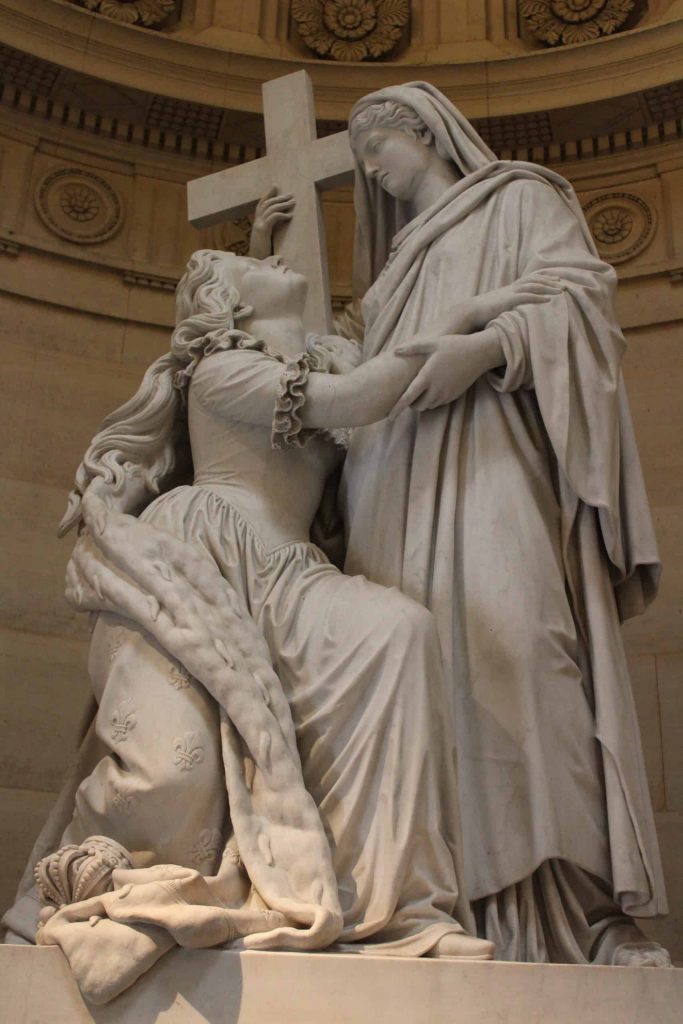 A photo of a statue of Marie Antoinette looking up a representation of religion.