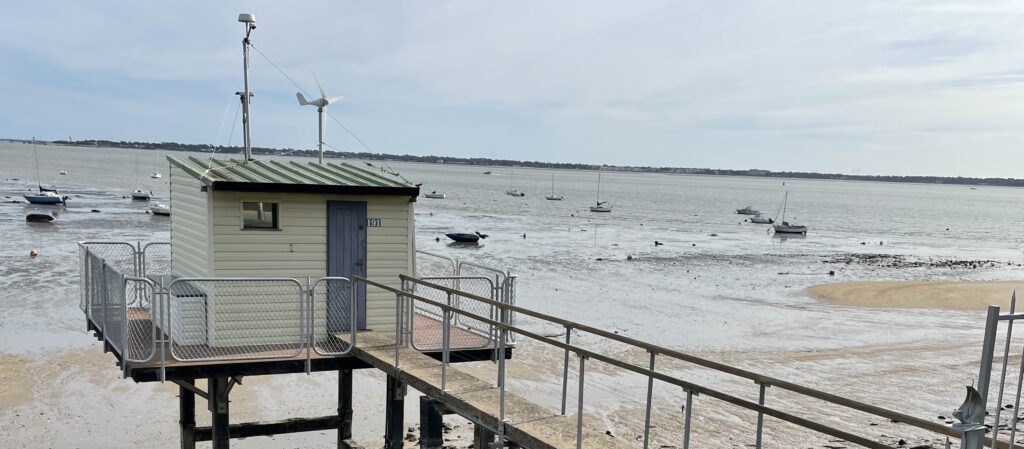 A photo of a fishing hut in Saint-Nazaire. It shows an elevated path leading out to the hut. The tide is out, so it is just sand underneath.