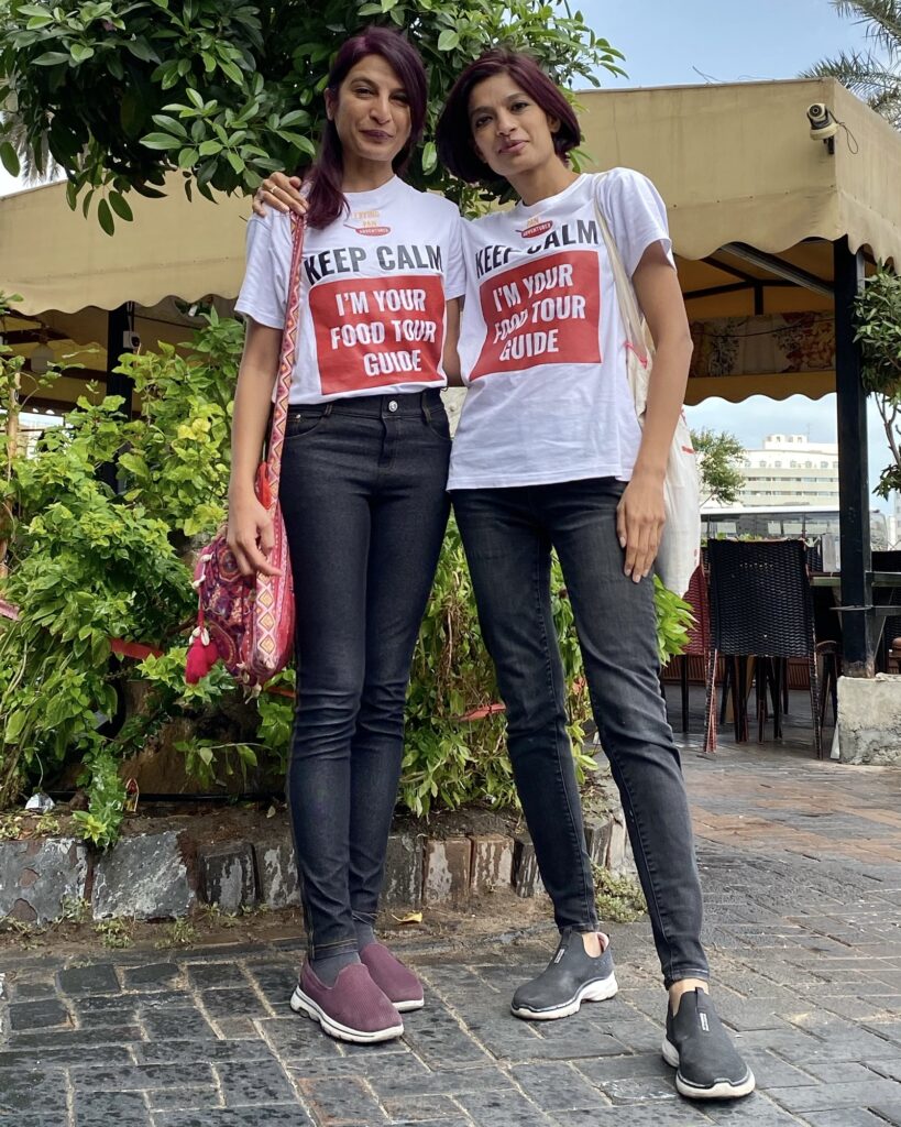 A photo of Arva and Farida, owners of Frying Pan Adventures. They are standing side by side, looking into the camera, wearing t-shirts that say Keep Calm, I'm Your Food Tour Guide.