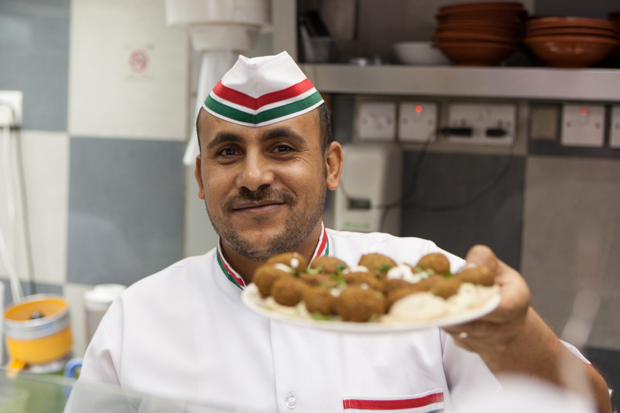 A photo of a food vendor looking into the camera with smile, holding out a plate of falafel.